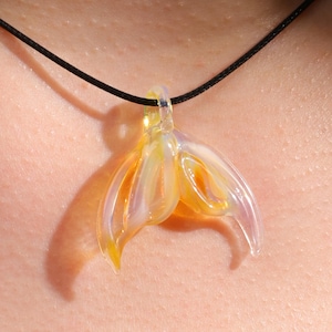 May include: A pendant necklace featuring a translucent, iridescent glass mermaid tail design. The pendant has yellow, orange, and purple hues and is suspended from a black cord. The necklace is a piece of jewelry.