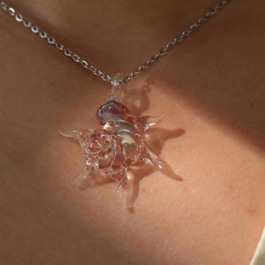 Pode incluir: Colar com pingente de vidro transparente com design de estrela. O pingente tem um centro em espiral e pontas radiantes, com toques de roxo e amarelo. A corrente do colar é prateada.