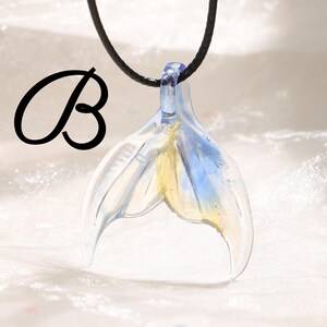 May include: A clear glass pendant in the shape of a mermaid's tail, with blue and yellow accents. The pendant is suspended from a black cord. The letter "B" is in the upper left corner.