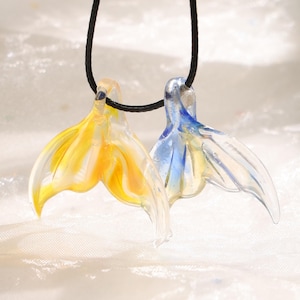 May include: Two glass mermaid tail pendants, one yellow and the other blue, hang from black cords. The pendants are translucent, with the yellow tail having orange accents and the blue tail having blue accents. The background is a soft, white fabric.