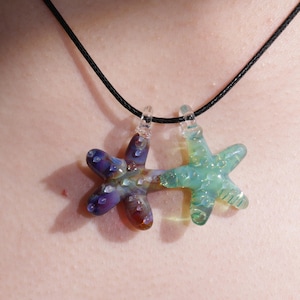 May include: A necklace with two glass starfish pendants. One is a mix of purple, blue, and brown, the other light green. Both have clear glass loops and are strung on a black cord. The pendants are likely handmade.