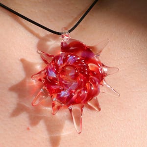 May include: A red and clear glass pendant necklace. The pendant is shaped like a sunburst with pointed rays. The center of the pendant has a spiral design with hints of purple and blue. The necklace has a black cord.