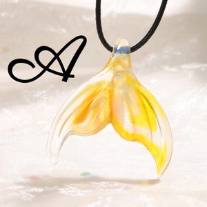 May include: A glass pendant shaped like a mermaid's tail, with yellow and clear coloring. The pendant is suspended from a black cord. The letter "A" is in black script in the upper left corner.
