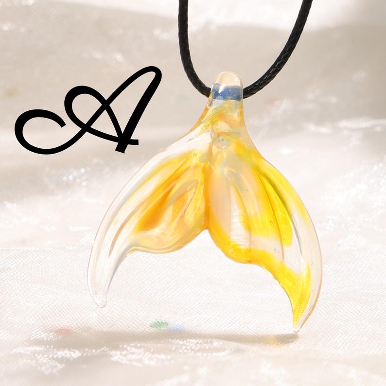 May include: A glass pendant shaped like a mermaid's tail, with yellow and clear coloring. The pendant is suspended from a black cord. The letter "A" is in black script in the upper left corner.