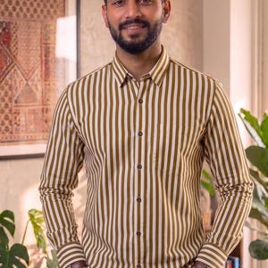Può includere: Camicia abbottonata a righe verticali verde oliva e bianche. La camicia ha una tasca sul petto e viene indossata con jeans blu. Le maniche sono lunghe e abbottonate ai polsini.