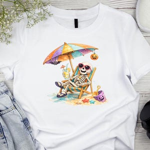 May include: A white t-shirt featuring a graphic of a skeleton wearing pink sunglasses, relaxing in a beach chair under a colorful striped umbrella. The skeleton holds a yellow drink, with a small bat, a jack-o'-lantern, and a purple bucket nearby.