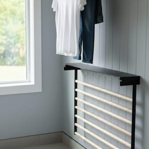 May include: A wall-mounted clothes drying rack with a black metal frame and natural wood dowels. A white t-shirt and blue jeans hang on the upper rack. The lower rack is empty. The rack is in a room with a window.