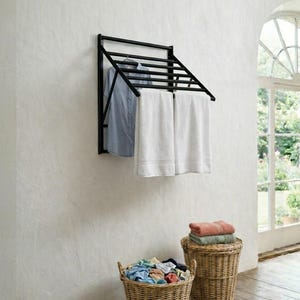 May include: A black metal wall-mounted drying rack with a blue shirt and two white towels hanging on it. Two wicker baskets are on the floor, one filled with clothes and the other with folded towels. The room has a rustic aesthetic.