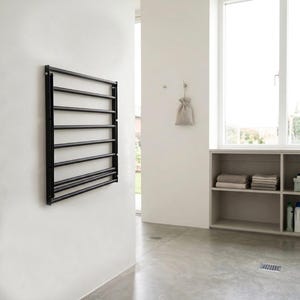 May include: A black, wall-mounted towel rack with seven horizontal bars. The rack is rectangular and mounted on a white wall. The room has a minimalist design with a window and a shelf with folded towels.