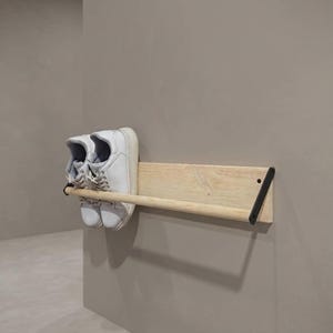 May include: A wall-mounted shoe rack made of light-coloured wood, holding a pair of white trainers. The rack has a minimalist design with black metal supports. The shoes are white.