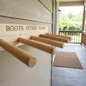 May include: Wooden boot rack mounted on a wall. The rack has five dowels for hanging boots and the text "BOOTS OUTSIDE PLEASE!" is engraved on the top. The rack is made of light-colored wood.