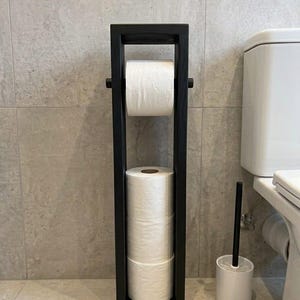 May include: A tall, rectangular black wooden toilet paper stand featuring a horizontal rod at the top for one roll and a vertical storage compartment at the bottom holding three stacked white toilet paper rolls, positioned next to a white toilet brush.