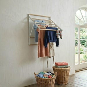 May include: A wall-mounted wooden clothes drying rack with clothes hanging on it. Below are two wicker baskets, one filled with folded clothes and the other with towels. The room has a rustic, natural aesthetic.