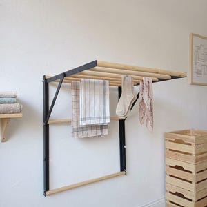 May include: A wall-mounted clothes drying rack with a black metal frame and wooden dowels. The rack holds a checked towel, socks, and a patterned scarf. A wooden shelf with folded towels and wooden crates are also visible.