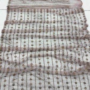 Puede incluir: Una tela de tul en color rosa empolvado con filas verticales de lentejuelas metálicas y pequeños motivos florales bordados. El material transparente muestra un patrón repetitivo de destellos dorados dispuestos en líneas rectas.