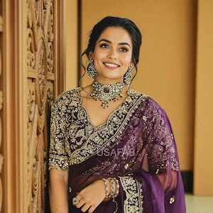 May include: A woman wearing a dark purple sari with gold embroidery and a matching blouse. The sari features intricate patterns and a sheer overlay. She is adorned with a necklace, earrings, and bracelets. The text "USAFAB" is visible on the sari.