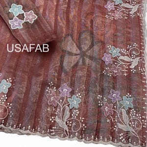 May include: A sheer, rust-colored fabric with vertical stripes and a subtle floral print. The edges feature scalloped borders and embroidered floral appliqués in light blue, lavender, and pink. The text USAFAB is printed on the fabric.