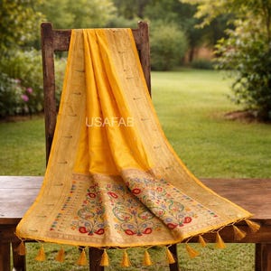 Puede incluir: Un textil amarillo vibrante con intrincados dise&ntilde;os florales y de cachemira, drapeado sobre una silla de madera. El textil presenta un borde detallado y borlas decorativas. El texto "USAFAB" es visible.