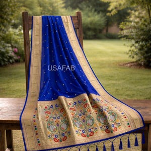 Puede incluir: Un sari azul real con bordes dorados y bordados florales intrincados. El sari est&aacute; drapeado sobre una silla y una mesa de madera, con el texto "USAFAB" visible. El sari tiene un borde decorativo con borlas.