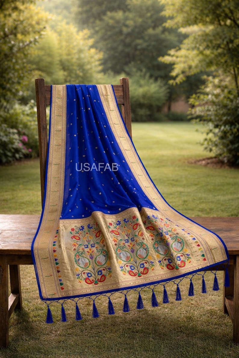 Puede incluir: Un sari azul real con bordes dorados y bordados florales intrincados. El sari est&aacute; drapeado sobre una silla y una mesa de madera, con el texto "USAFAB" visible. El sari tiene un borde decorativo con borlas.