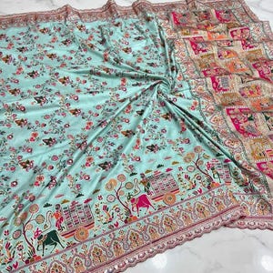 May include: A light blue fabric textile featuring a dense floral pattern in pink, red, and green. The border displays a detailed grid design with colourful illustrations of figures, carts, and animals, finished with a scalloped, embroidered edge.