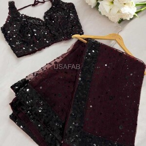 May include: A burgundy sequined blouse and matching saree with a black sequined border. The saree is draped over a wooden hanger. The text "USAFAB" is visible on the saree. A bouquet of white flowers is in the background.