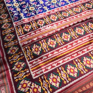 May include: A detailed shot of a textile with a vibrant floral pattern. The fabric features a deep blue background with yellow, pink, and white floral motifs. Borders in red, yellow, and white frame the design, creating a rich, textured appearance.