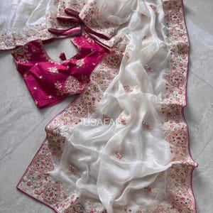 Peut inclure: Un sari blanc avec des broderies complexes et un chemisier fuchsia assorti. Le sari présente des motifs floraux détaillés et une bordure délicate. Le chemisier est d'un fuchsia vif, complétant le design du sari. L'ensemble est élégant et traditionnel.