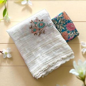 May include: White fabric with floral embroidery in blue, pink, and peach. The fabric has a scalloped edge and is paired with a patterned teal fabric with pink and orange flowers. The word "SAFA" is visible on the white fabric.