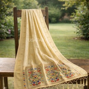 Puede incluir: Textil color crema con intrincados bordados con dise&ntilde;os florales y de cachemira en rojo, azul, amarillo y verde. El textil est&aacute; drapeado sobre una silla y una mesa de madera, con borlas en el borde inferior. El texto "USAFAB" es visible.