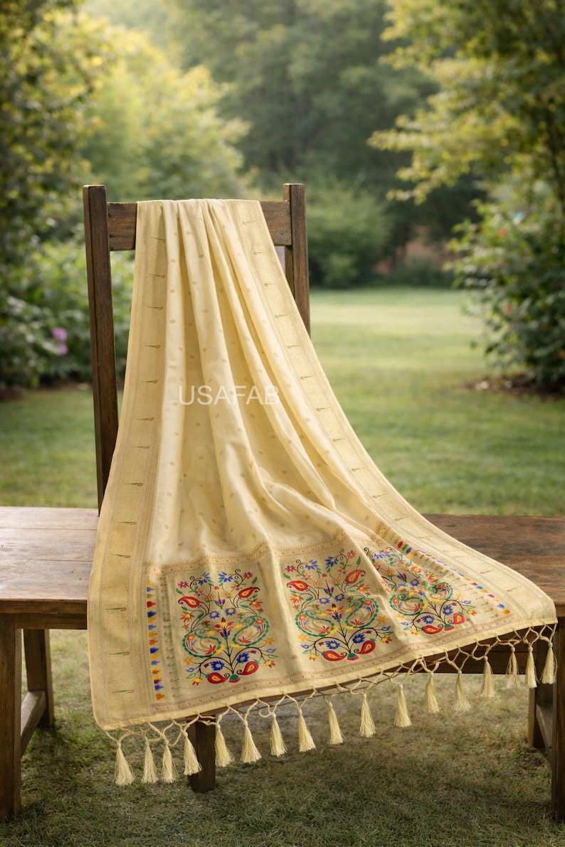 Puede incluir: Textil color crema con intrincados bordados con dise&ntilde;os florales y de cachemira en rojo, azul, amarillo y verde. El textil est&aacute; drapeado sobre una silla y una mesa de madera, con borlas en el borde inferior. El texto "USAFAB" es visible.