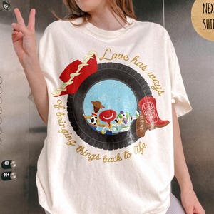 May include: An ivory t-shirt featuring a graphic of a black tire framing a scene with cartoon characters, a red cowboy hat, and a red cowboy boot. The text reads, Love has ways of bringing things back to life, in a cursive gold font.
