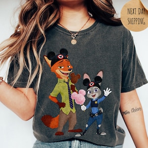 May include: Dark gray t-shirt featuring a cartoon illustration of Nick Wilde and Judy Hopps from Zootopia, each wearing Mickey Mouse ears. Nick holds a chocolate treat, and Judy holds a pink cotton candy. The word "PEPPER" is at the bottom.