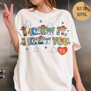 May include: An ivory short-sleeve t-shirt featuring a graphic print with the text I KNEW IT, I KNEW YOU and TS inside a red heart. The letters are decorated with patterns inspired by toy characters, including cowboy hats, space ranger suits, and stars.