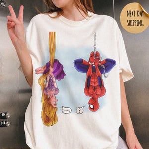 Op de afbeelding: Een ivoorkleurig t-shirt met een grafische print van een langharig personage dat aan haar haar hangt en een rood-blauwe superheld die aan een web hangt. Het personage zegt O HAI en de superheld heeft een vraagteken in een tekstballon.
