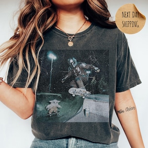 May include: Dark gray t-shirt featuring a graphic of The Mandalorian and Grogu skateboarding in a skate park. The image has a vintage, distressed look. The shirt has a crew neck and short sleeves. The word "PEPPER" is printed at the bottom.