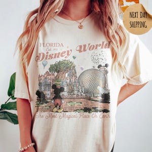 May include: Ivory-colored t-shirt featuring a vintage-style graphic of Disney World in Florida. The design includes Mickey Mouse, a castle, Spaceship Earth, and the text "Florida Disney World Est. 1971" and "The Most Magical Place On Earth."