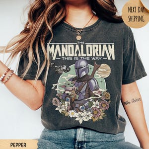 May include: Pepper-colored t-shirt featuring a graphic of The Mandalorian and Grogu, with the text "The Mandalorian - This is the Way." The design includes a space scene with flowers. The shirt has a vintage, worn look.