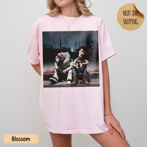 Pode incluir: Camiseta rosa claro com uma estampa de Buzz Lightyear e Woody de Toy Story sentados em uma cal&ccedil;ada. A imagem &eacute; definida em um fundo escuro e urbano. A palavra "Blossom" &eacute; impressa na parte inferior.