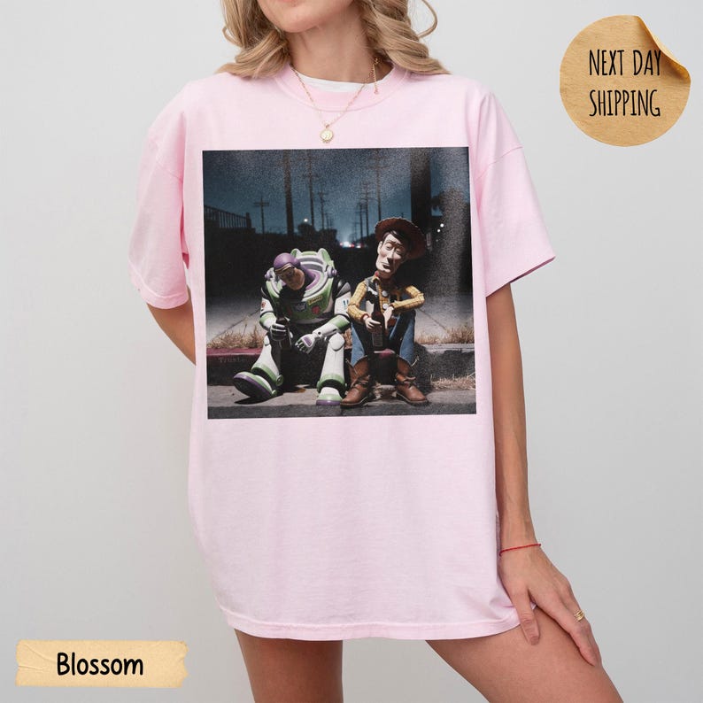 Pode incluir: Camiseta rosa claro com uma estampa de Buzz Lightyear e Woody de Toy Story sentados em uma cal&ccedil;ada. A imagem &eacute; definida em um fundo escuro e urbano. A palavra "Blossom" &eacute; impressa na parte inferior.