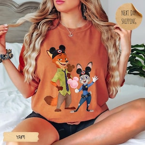 May include: Orange-colored t-shirt featuring a cartoon illustration of Nick Wilde and Judy Hopps from Zootopia. Nick is wearing a green shirt and brown pants, while Judy is in a police uniform. The shirt has the text "YAM" at the bottom.