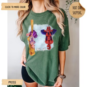 Op de afbeelding: Een mosgroen T-shirt met korte mouwen en een grafische print van een vrouwelijk personage met lang blond haar dat aan een vlecht hangt en een rood-blauw superheldpersonage dat aan een web hangt. De tekst O HAI en een vraagteken zijn afgedrukt.