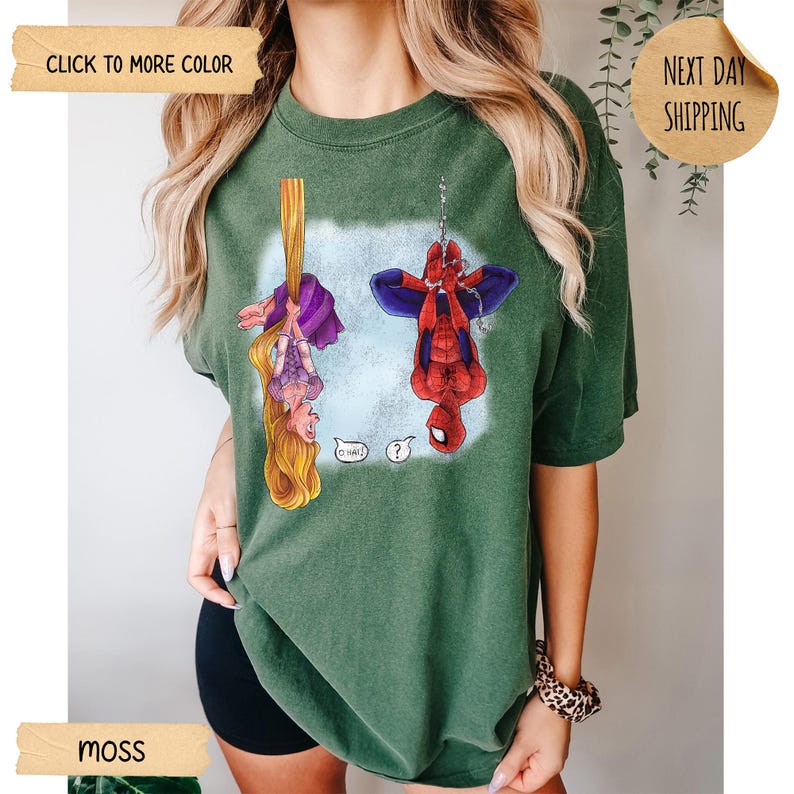 Op de afbeelding: Een mosgroen T-shirt met korte mouwen en een grafische print van een vrouwelijk personage met lang blond haar dat aan een vlecht hangt en een rood-blauw superheldpersonage dat aan een web hangt. De tekst O HAI en een vraagteken zijn afgedrukt.