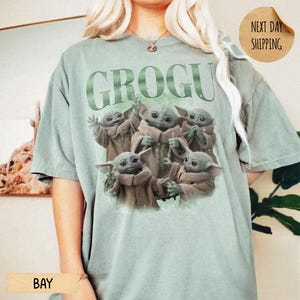 May include: A sage green short-sleeve t-shirt featuring the text GROGU in large green letters above a graphic illustration of five small green creatures with large ears and eyes, each wearing a tan robe, arranged in a group on the front of the shirt.