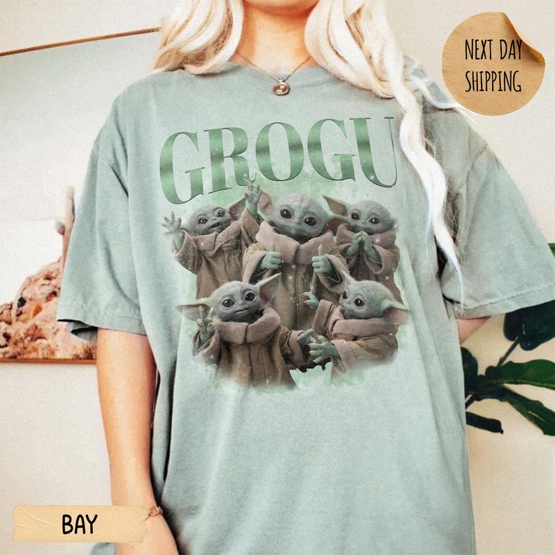 May include: A sage green short-sleeve t-shirt featuring the text GROGU in large green letters above a graphic illustration of five small green creatures with large ears and eyes, each wearing a tan robe, arranged in a group on the front of the shirt.