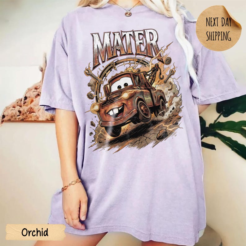 May include: Orchid purple t-shirt with a graphic of Mater from the Cars film. The design includes the word "MATER" in a distressed font above the character. The image shows Mater kicking up dirt and has a "Next Day Shipping" sticker.