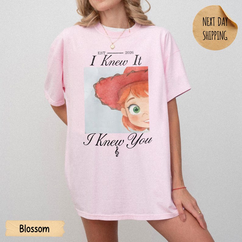 May include: A light pink t-shirt featuring a graphic of a cartoon character wearing a red hat. The text above the graphic reads "EST 2026 I Knew It" and the text below reads "I Knew You" with a small musical treble clef symbol centered underneath.