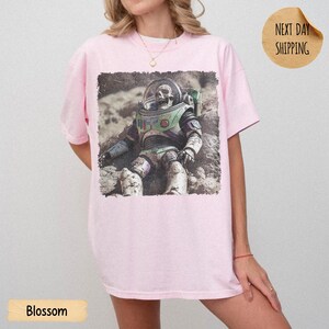 May include: A light pink t-shirt featuring a graphic print of a weathered, skeletal astronaut figure in a space suit sitting on a textured, rocky surface. The astronaut suit displays muted green, purple, and white tones with a clear helmet visor.