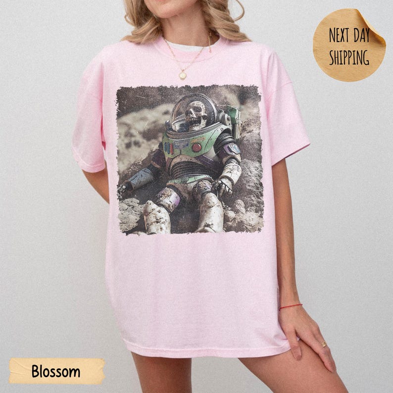 May include: A light pink t-shirt featuring a graphic print of a weathered, skeletal astronaut figure in a space suit sitting on a textured, rocky surface. The astronaut suit displays muted green, purple, and white tones with a clear helmet visor.