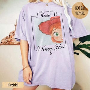 May include: A light purple short-sleeve t-shirt featuring a graphic print of a cartoon character with red hair and a red hat. The text reads EST 2026 I Knew It I Knew You with a musical note symbol. The design is framed by a thin black rectangular border.